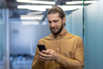 A young professional man with a beard, engaged in using his smartphone, stands in a contemporary...