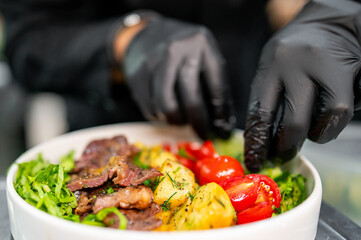 Person in black gloves preparing a vibrant salad with grilled meat, fresh vegetables, and greens. Perfect for culinary and healthy lifestyle concepts