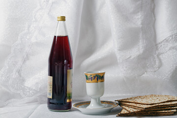 Bottle of red wine, a goblet, and matzah bread against a white lace curtain backdrop. The goblet has intricate designs. The matzo is stacked neatly beside the goblet, Jewish Passover Seder meal.