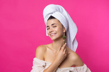 A beautiful cheerful attractive girl with a towel on her head holds a cosmetic roller near her face against a pink background.