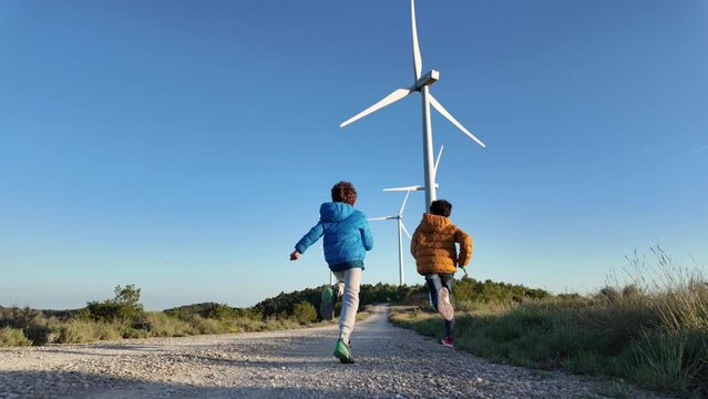 Two kids, aged 8 and 6, run amidst a field with wind turbines. Emphasizing sustainability, environmental care, renewable energy, and energy freedom.