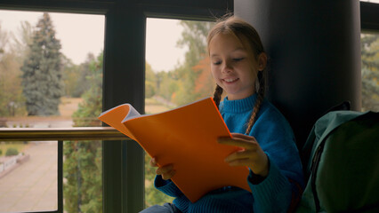 Little girl sitting school hall reading textbook prepare exam task homework lesson indoors...