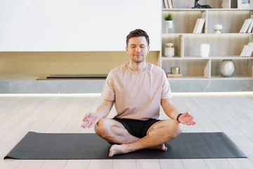  Man seated in lotus position meditates on a yoga mat, cultivating inner peace in a minimalist, bright room. © muse studio