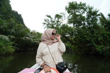 A female model wearing a hijab is sitting on a wooden boat sailing around a river with many mangrove forests on its side - Powered by Adobe