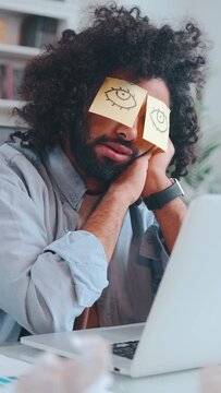 Young lazy funny Arabian man office worker with paper stickers on eyes sleeps working and pretends so as not to be fired or fined sits at workplace at table with laptop. Overwork, overload, burnout