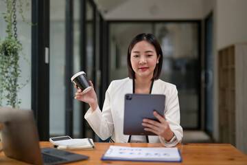 Smiling professional businesswoman holding a tablet in a contemporary office setting, relax with coffee.