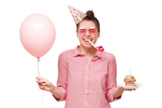 Young happy girl in glasses and birthday hat, blowing whistle, holding pink balloon and cupcake, laughing and enjoying party - Powered by Adobe