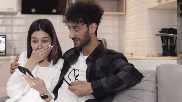 Young woman showing something to her partner on her smartphone and becoming rejoice and happy while they are sitting together on a couch in a cozy kitchen at home