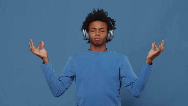 Young black guy in headphones trying to relax and starting to meditate on a blue background in a studio. Man connecting the thumb and forefinger on both hands and putting them to the sides