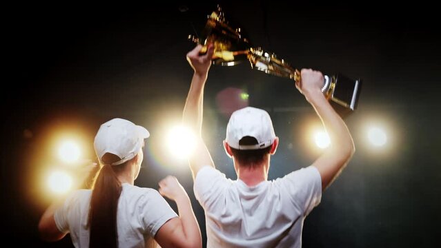 Two competition winners joy for sport success and raise trophy up standing on stage back view. Team celebrating victory together, achievement motivation
