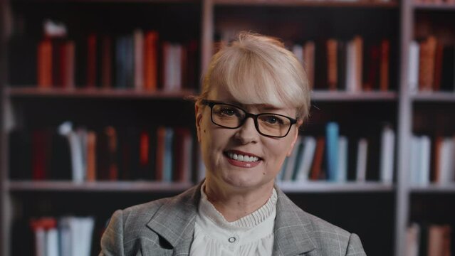 Portrait of a mature caucasian woman in glasses, standing, looking at the camera, and smiling with a backdrop of bookshelves with many books