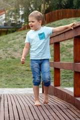 A little barefoot boy in blue clothes is walking on a wooden bridge in a city park, holding onto the railing