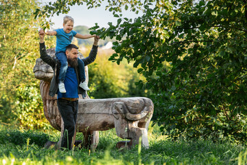Father and son friendship, the father carries the child on his shoulders. Beautiful summer nature, a wooden lion-shaped bench for rest nearby.