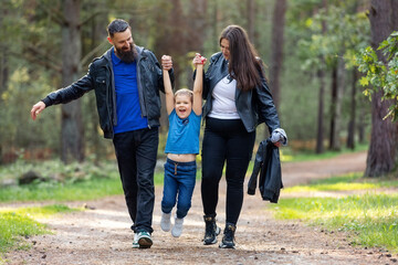 Family walk in the park. Parents lift their child by the arms. The little boy is very happy. A beautiful summer evening in a pine forest