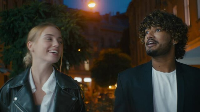 Happy couple European woman Indian man friends walking street city center talking outside evening looking smile laugh together stroll fun meeting girl guy pointing show conversation chatting weekend