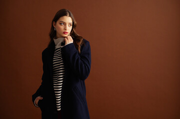 Fashionable confident woman wearing stylish striped turtleneck sweater, classic navy blue woolen coat, posing on brown background. Studio fashion portrait. Copy, empty, blank space for text 