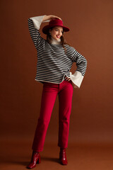 Fashionable happy smiling woman wearing stylish striped oversize turtleneck sweater, red jeans, hat, patent leather ankle boots, posing on brown background. Full-length studio fashion portrait - 790938821