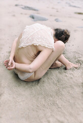 woman with fine lingerie and embroidered vintage cape curled up on herself on the sand on a beach on the Asturian coast