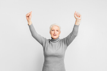 Joyful senior lady with arms raised on grey background