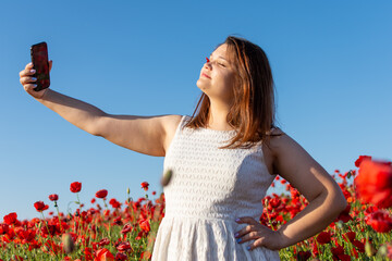 Woman in white dress smiles, taking selfie in red flower field under sunny sky