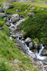 A small waterfall flows through rocks and grass in mountainous terrain