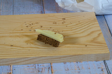 "Close-up of half a slice of rye bread with a piece of semi-cured cheese on top on a light wooden cutting board, all on a wooden table with faded grayish-blue painted old boards."