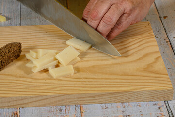 "On a light wooden cutting board, a hand and a knife are seen cutting pieces of semi-cured Manchego cheese, all on a table of faded rustic planks painted with a grayish-blue hue."