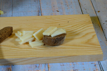 "Close-up of half a slice of rye bread with a piece of semi-cured cheese on top on a light wooden cutting board, all on a wooden table with faded grayish-blue painted old boards."