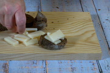 A slice of rye bread with some pieces of semi-cured cheese on top and beside it, on a light wooden cutting board, all placed on a wooden table with faded grayish-blue painted old boards.hand of a man 