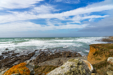 Fototapeta na wymiar Sur le littoral atlantique de la baie d'Audierne, ciel bleu, nuages blancs, rochers lichens orange, sillons rocheux, écume de mer.