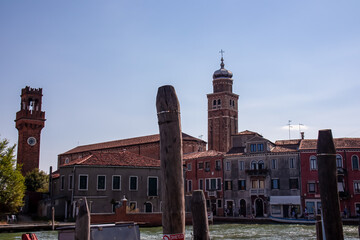 Panoramic view of bell tower of Chieso di San Pietro Martire next to water channel on Murano island in city of Venice, Veneto, Italy, Europe. Venetian architectural landmarks and wooden timber piles - obrazy, fototapety, plakaty
