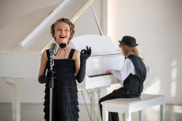 A young girl in a black dress sings into a microphone while a boy plays the piano.