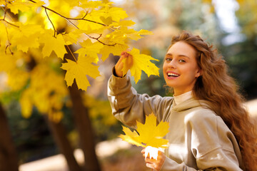 Smiling young woman enjoys the autumn weather at the maple tree with the yellow leaves at sunset