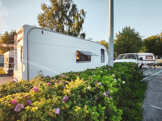 A serene and charming sight of a quaint small white retro travel trailer nestled among colorful flowers and green bushes under a clear blue sky, offering a tranquil setting for a relaxing getaway.