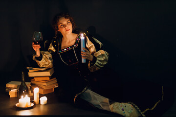 Young adult woman dressed in a medieval dress sitting near ancient books and candles with bottle of red wine and holding a glass of wine in darkness