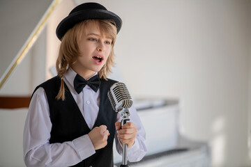 A young boy in a black vest and hat singing into a microphone.