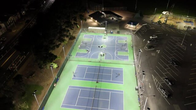 Drone flying at night above illuminated basket and tennis pitches with people playing