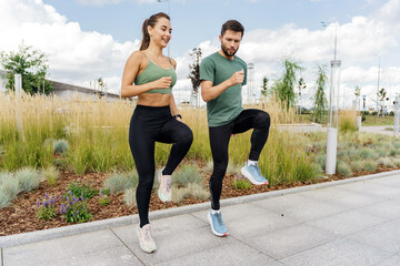 Energetic couple in sportswear perform dynamic high-knee exercises in an urban green space.