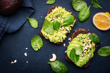 Avocado toasts with spinach and cashew nuts sprinkled with sesame seeds on black table background,...