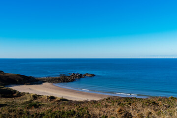 Falaise et pointe rocheuse encadrent la plage de sable de la Fosse, Côtes d'Armor, sous un ciel bleu azur, créant un paysage marin pittoresque.