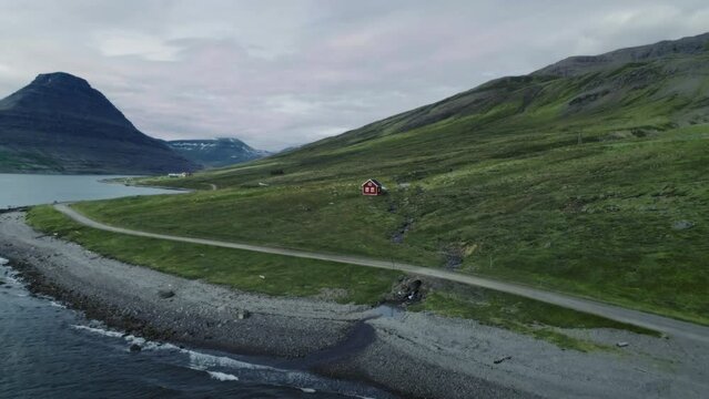 Aerial drone shot of adorable cute little wooden traditional red house on green hill in rural Iceland. Gravel road along the coastline of rugged Westfjords. Cinematic rough icelandic climate 