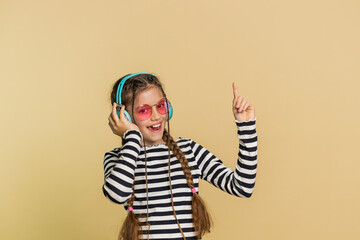 Happy smiling preteen child girl in headphones listening music dancing disco fooling around having fun expressive gesticulating hands relaxing on party making funny moves. Kid on beige background