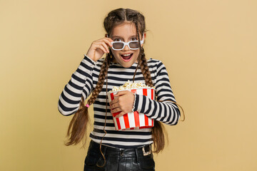 Excited amazed young preteen child girl kid eating popcorn and watching movie film wearing 3D cinema glasses, enjoying snacks, online content. Teenager Caucasian children isolated on beige background