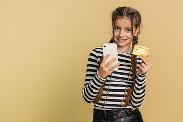 Happy preteen child girl kid using credit bank card and smartphone while transferring spending money, purchases online shopping, ordering food delivery. Young children isolated on beige background