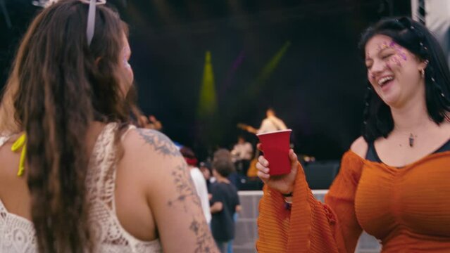 Two female friends wearing glitter and drinks having fun dancing at outdoor summer music festival - shot in slow motion 
