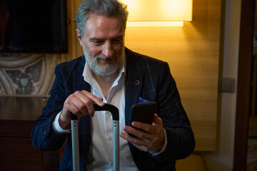 Adult caucasian businessman with suitcase watching smartphone in hotel room