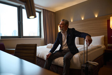 Mature businessman with suitcase sitting on bed and looking away in hotel room