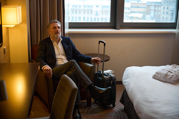 Thoughtful caucasian businessman with suitcase sitting in armchair in hotel room