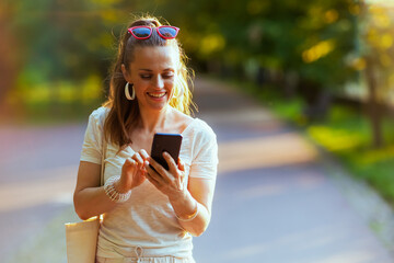 smiling trendy woman in shirt using smartphone and walking