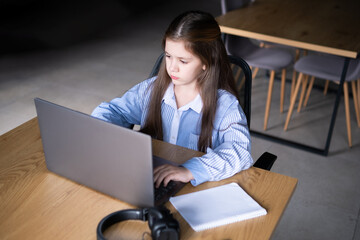 A cute small girl sitting alone at a table looking in a laptop screen doing her distance education...